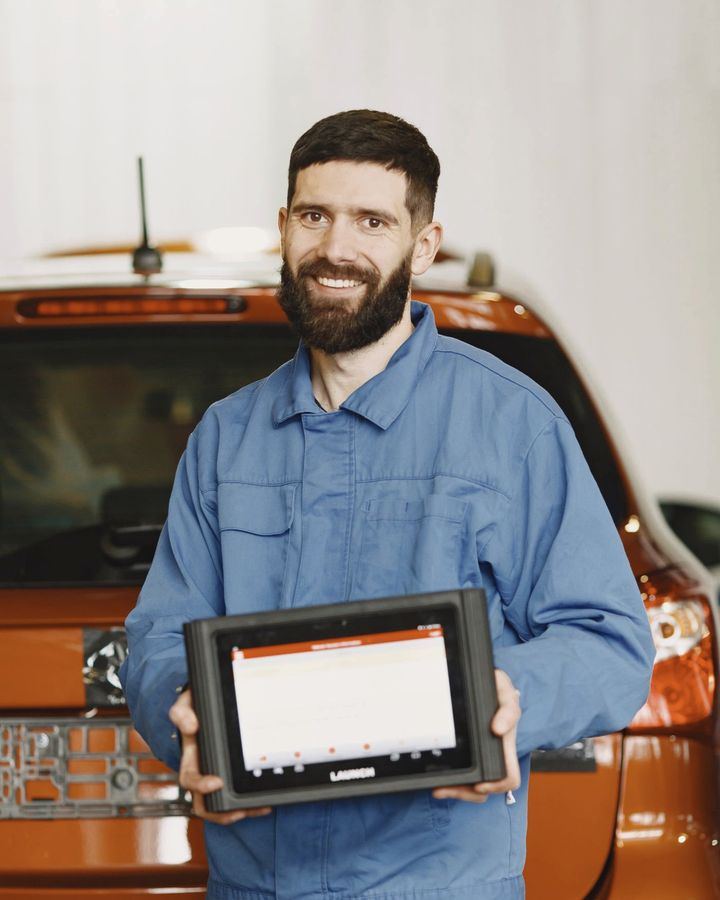 An auto repair technician in coveralls holding a diagnostic tablet beside a car.