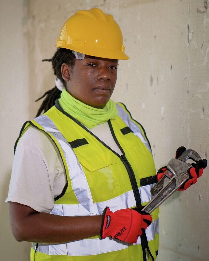 A tradesperson in a hi-vis vest and hard hat holding a pipe wrench on a job site.