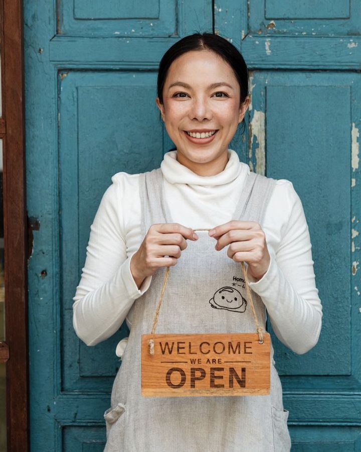 A shop owner in an apron holding a hand-lettered open sign in her doorway.